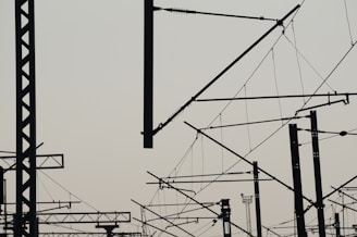 A complex network of silhouetted train electrical lines and poles with a light sky background, creating an intricate lattice of intersecting shapes against a pale backdrop.