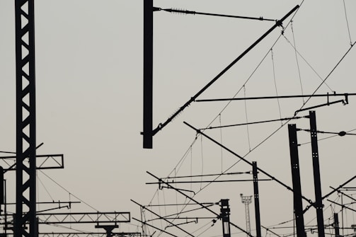 A complex network of silhouetted train electrical lines and poles with a light sky background, creating an intricate lattice of intersecting shapes against a pale backdrop.