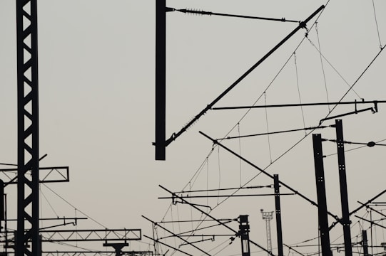 A complex network of silhouetted train electrical lines and poles with a light sky background, creating an intricate lattice of intersecting shapes against a pale backdrop.