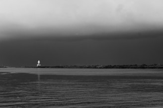 A black and white landscape featuring a body of water in the foreground and a distant shoreline. A lighthouse stands prominently on the shoreline, with a moody, overcast sky above.