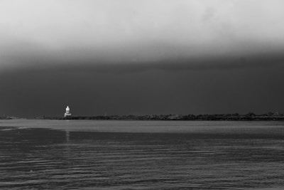 A black and white landscape featuring a body of water in the foreground and a distant shoreline. A lighthouse stands prominently on the shoreline, with a moody, overcast sky above.