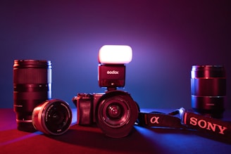 A collection of photography equipment including a Sony camera with a lens attached, an external flash mounted on top, and multiple camera lenses placed around the main camera. The setup is illuminated by a mix of red and blue lighting, creating a dramatic effect.