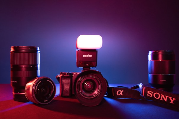 A collection of photography equipment including a Sony camera with a lens attached, an external flash mounted on top, and multiple camera lenses placed around the main camera. The setup is illuminated by a mix of red and blue lighting, creating a dramatic effect.