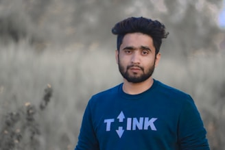 A man with short, dark hair and a beard is standing outdoors. He is wearing a dark blue shirt with the word 'THINK' printed on it in white letters. The background features out-of-focus greenery, suggesting a natural setting.