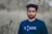 A man with short, dark hair and a beard is standing outdoors. He is wearing a dark blue shirt with the word 'THINK' printed on it in white letters. The background features out-of-focus greenery, suggesting a natural setting.
