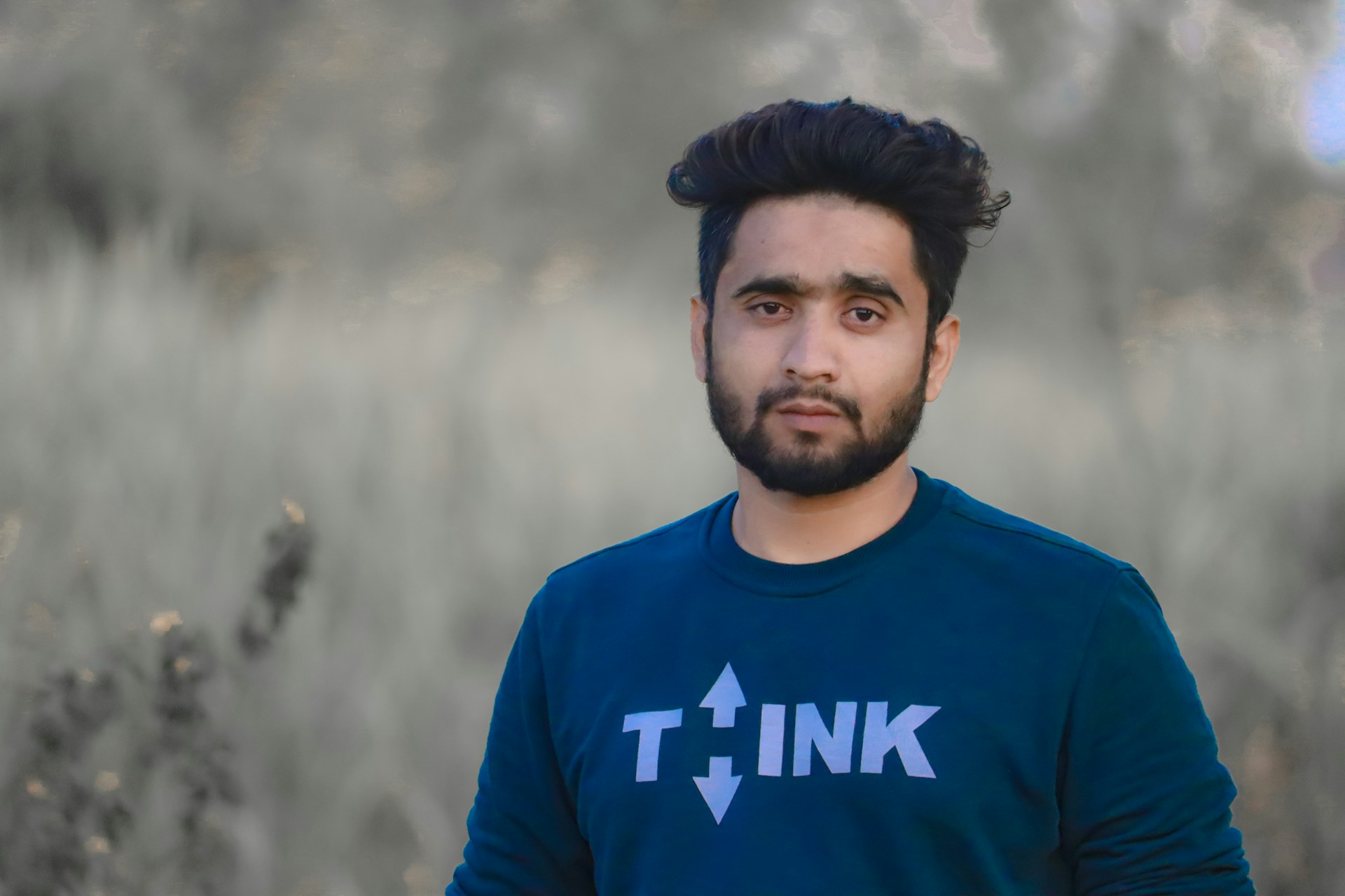 A man with short, dark hair and a beard is standing outdoors. He is wearing a dark blue shirt with the word 'THINK' printed on it in white letters. The background features out-of-focus greenery, suggesting a natural setting.