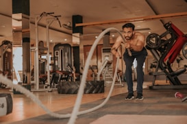 A man is engaged in an intense workout session in a gym environment. He is shirtless and performing battle rope exercises with visible determination. The setting includes various gym equipment like a leg press machine, dumbbells, a large tire, and other exercise machines. The room has a warm, natural lighting that highlights the man's focus and physical exertion.