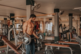 A muscular man is lifting a barbell in a well-equipped gym. He appears focused and determined while performing the exercise. The gym includes various workout machines and benches, and it's well-lit with natural and artificial light.