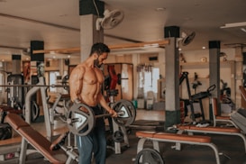 A muscular man is lifting a barbell in a well-equipped gym. He appears focused and determined while performing the exercise. The gym includes various workout machines and benches, and it's well-lit with natural and artificial light.