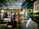A tailor shop window display featuring a plaid suit on a mannequin, with elegant gold lettering that reads 'Kings Inch Tailor Est. 1943.' Inside the shop, there are neatly organized racks of shirts, jackets, and shoes. The interior has a vintage style with dark wooden shelves and green walls. A framed photograph of a man holding a child's hand adds a personal touch.