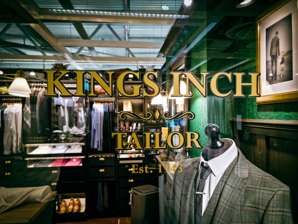 A tailor shop window display featuring a plaid suit on a mannequin, with elegant gold lettering that reads 'Kings Inch Tailor Est. 1943.' Inside the shop, there are neatly organized racks of shirts, jackets, and shoes. The interior has a vintage style with dark wooden shelves and green walls. A framed photograph of a man holding a child's hand adds a personal touch.