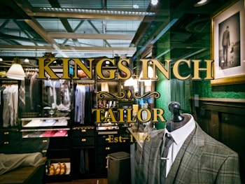 A tailor shop window display featuring a plaid suit on a mannequin, with elegant gold lettering that reads 'Kings Inch Tailor Est. 1943.' Inside the shop, there are neatly organized racks of shirts, jackets, and shoes. The interior has a vintage style with dark wooden shelves and green walls. A framed photograph of a man holding a child's hand adds a personal touch.