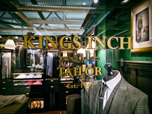 A tailor shop window display featuring a plaid suit on a mannequin, with elegant gold lettering that reads 'Kings Inch Tailor Est. 1943.' Inside the shop, there are neatly organized racks of shirts, jackets, and shoes. The interior has a vintage style with dark wooden shelves and green walls. A framed photograph of a man holding a child's hand adds a personal touch.