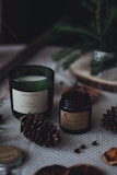 Aromatic candles in green and dark brown jars are placed on a dotted fabric surface. Pine cones and other natural elements surround the candles, contributing to a cozy, forest-like setting. A small evergreen branch is also visible in the background, adding to the rustic feel.