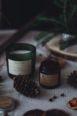 Aromatic candles in green and dark brown jars are placed on a dotted fabric surface. Pine cones and other natural elements surround the candles, contributing to a cozy, forest-like setting. A small evergreen branch is also visible in the background, adding to the rustic feel.