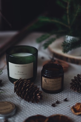 Aromatic candles in green and dark brown jars are placed on a dotted fabric surface. Pine cones and other natural elements surround the candles, contributing to a cozy, forest-like setting. A small evergreen branch is also visible in the background, adding to the rustic feel.