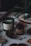 Aromatic candles in green and dark brown jars are placed on a dotted fabric surface. Pine cones and other natural elements surround the candles, contributing to a cozy, forest-like setting. A small evergreen branch is also visible in the background, adding to the rustic feel.
