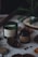 Aromatic candles in green and dark brown jars are placed on a dotted fabric surface. Pine cones and other natural elements surround the candles, contributing to a cozy, forest-like setting. A small evergreen branch is also visible in the background, adding to the rustic feel.