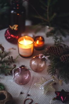 A cozy and festive atmosphere with candles illuminating a table decorated with Christmas ornaments, pine cones, and evergreen branches. Two candles are lit, with warm light reflecting off shiny, round ornaments. There is a star-shaped decoration adorned with beads, set on a textured tablecloth.