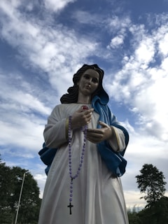 A statue of a religious figure is depicted, wearing a white robe with a blue shawl, holding a rosary with purple beads and a cross. The background consists of a partly cloudy sky, and surrounding the statue are trees.