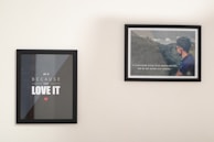 Two framed posters are hanging on a plain wall. The left poster features motivational text in bold white letters on a black background, with a small red heart below. The right poster displays an image of a man in a blue shirt facing a scenic landscape overlaid with another motivational quote in white text.