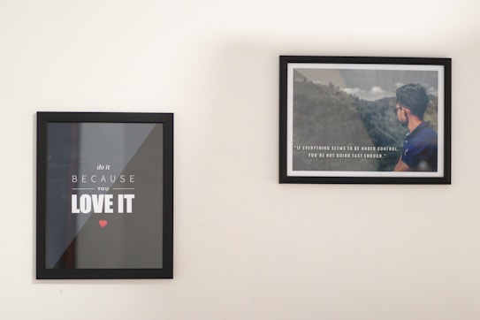 Two framed posters are hanging on a plain wall. The left poster features motivational text in bold white letters on a black background, with a small red heart below. The right poster displays an image of a man in a blue shirt facing a scenic landscape overlaid with another motivational quote in white text.