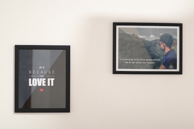 Two framed posters are hanging on a plain wall. The left poster features motivational text in bold white letters on a black background, with a small red heart below. The right poster displays an image of a man in a blue shirt facing a scenic landscape overlaid with another motivational quote in white text.