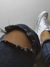 A close-up view of a slightly bent denim-clad leg with a large tear over the knee area, revealing skin. The fabric has frayed edges around the tear. Next to the leg is a white sneaker with visible shoelaces on a light-colored surface.