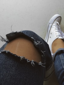 A close-up view of a slightly bent denim-clad leg with a large tear over the knee area, revealing skin. The fabric has frayed edges around the tear. Next to the leg is a white sneaker with visible shoelaces on a light-colored surface.