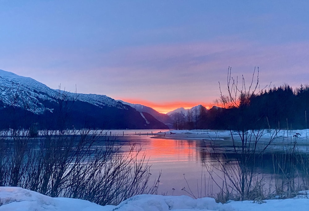 a lake surrounded by snow covered mountains at sunset