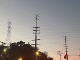 A series of utility poles and electrical wires are silhouetted against a clear sky during twilight. In the background, there is a steel transmission tower, and the foreground includes some streetlights and traffic lights.