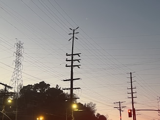A series of utility poles and electrical wires are silhouetted against a clear sky during twilight. In the background, there is a steel transmission tower, and the foreground includes some streetlights and traffic lights.