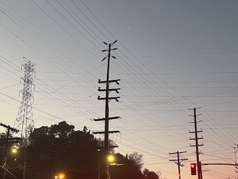 A series of utility poles and electrical wires are silhouetted against a clear sky during twilight. In the background, there is a steel transmission tower, and the foreground includes some streetlights and traffic lights.
