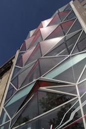 The image depicts a modern building facade with an intricate geometric glass design. The glass panels are arranged in triangular shapes and have varying shades of red, silver, and clear glass. The building structure shows a reflection of the trees and sky, indicating the surroundings.