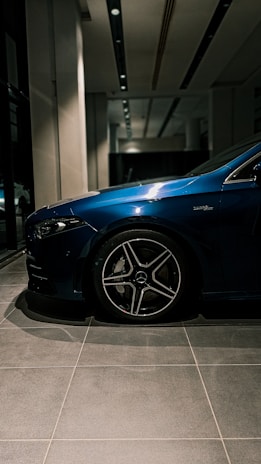 A close-up view of a blue car parked indoors, with focus on the wheel and part of the side body. The setting is a well-lit showroom with tiled flooring and a modern interior design.