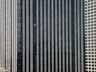 A person suspended midway up a tall skyscraper with dark vertical stripes between lighter concrete-colored stripes. The building features reflective windows, and the person appears to be a window washer or maintenance worker.