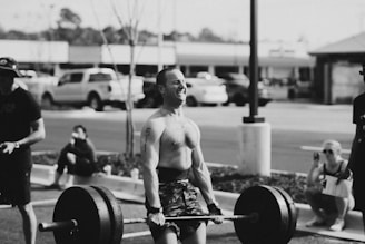 A person is lifting a heavy barbell outdoors, surrounded by a small group of spectators. The environment appears to be a parking lot, and the scene conveys physical exertion and focus.