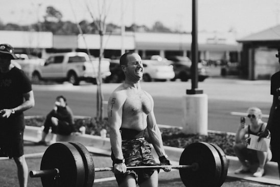 A person is lifting a heavy barbell outdoors, surrounded by a small group of spectators. The environment appears to be a parking lot, and the scene conveys physical exertion and focus.