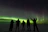 A group of happy travelers watching vivid Northern Lights dance over a snowy Icelandic landscape.