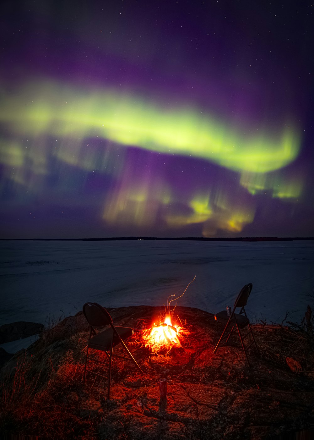 a campfire with two chairs and an aurora bore in the background