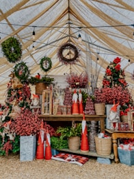 A festive display under a canopy with various Christmas decorations, including wreaths, small gnome figures, and red berries. The setting includes rustic elements like wooden tables and woven baskets, adorned with greenery and bright red accents.