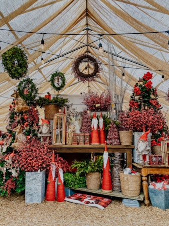 A festive display under a canopy with various Christmas decorations, including wreaths, small gnome figures, and red berries. The setting includes rustic elements like wooden tables and woven baskets, adorned with greenery and bright red accents.