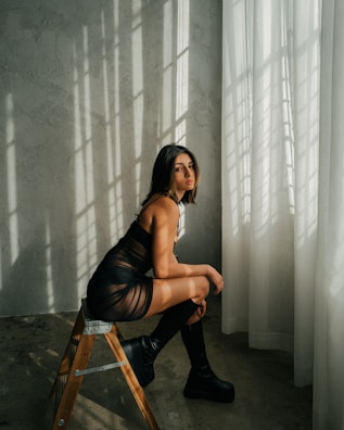 A woman sits on a wooden ladder stool in a room with soft, natural light filtering through sheer curtains. The sunlight casts intricate shadows on the wall and floor. She is wearing a sheer black outfit and high platform boots.
