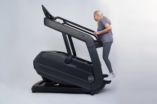 An older adult woman with long gray hair exercises on a large, dark-colored treadmill, dressed in gray activewear and white sneakers.