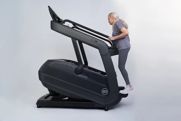 An older adult woman with long gray hair exercises on a large, dark-colored treadmill, dressed in gray activewear and white sneakers.