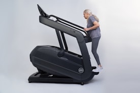 An older adult woman with long gray hair exercises on a large, dark-colored treadmill, dressed in gray activewear and white sneakers.