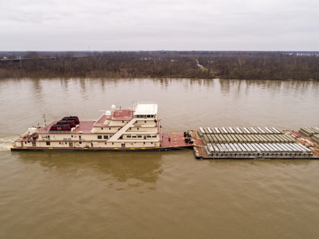 A large barge is transporting numerous cargo containers along a wide river. The water appears calm, and the surrounding landscape is flat with a line of trees on the horizon under an overcast sky.