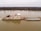 A large barge is transporting numerous cargo containers along a wide river. The water appears calm, and the surrounding landscape is flat with a line of trees on the horizon under an overcast sky.