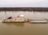 A large barge is transporting numerous cargo containers along a wide river. The water appears calm, and the surrounding landscape is flat with a line of trees on the horizon under an overcast sky.