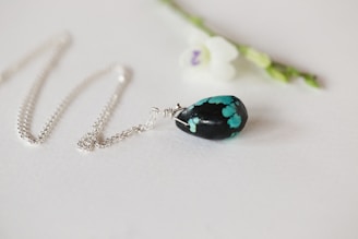 A delicate silver chain necklace with a pendant featuring a black stone with turquoise patterns. The necklace is elegantly laid out on a light surface next to a small white flower with a purple center and green stem.